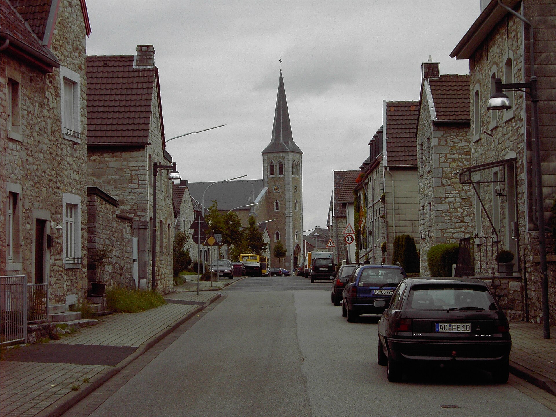 Die Straße Alt-Breinig mit Bruchsteinhäusern zu beiden Seiten, im Hintergrund der Turm von St. Barbara.