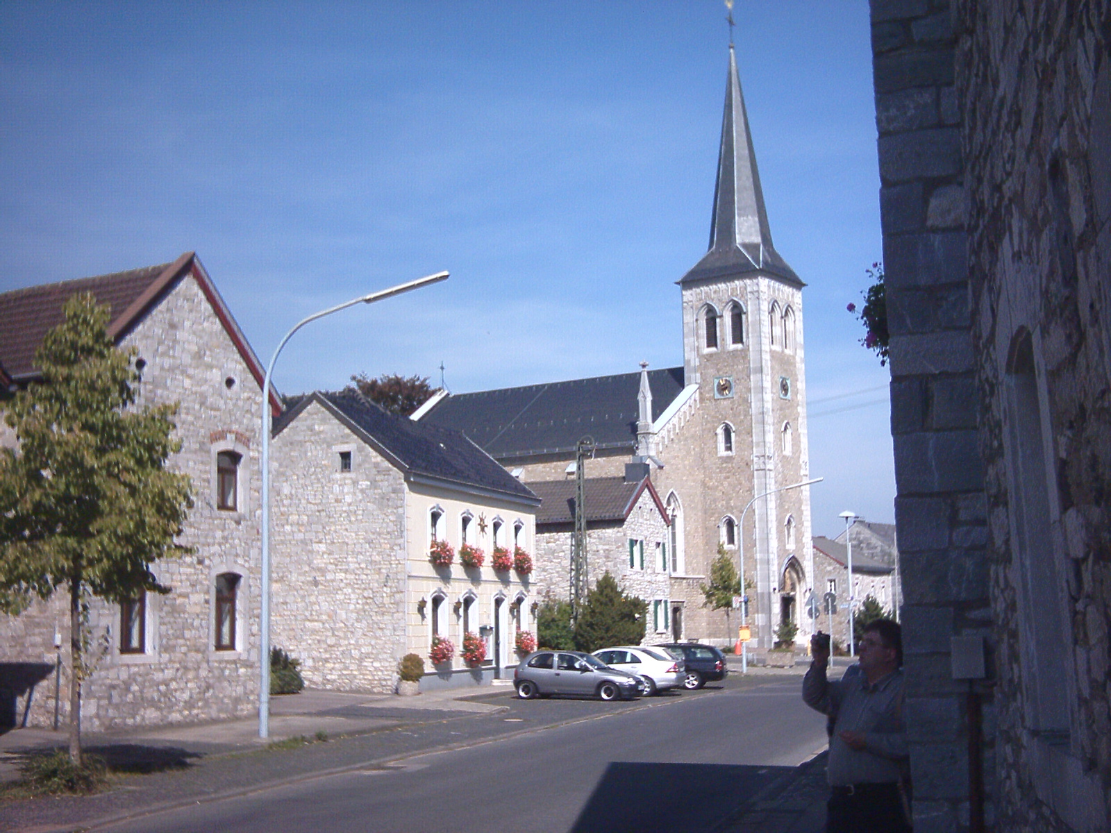 Die Pfarrkirche St. Barbara in Breinig mit spitzem Turm, davor Häuser aus hellem Kalkbruchstein.