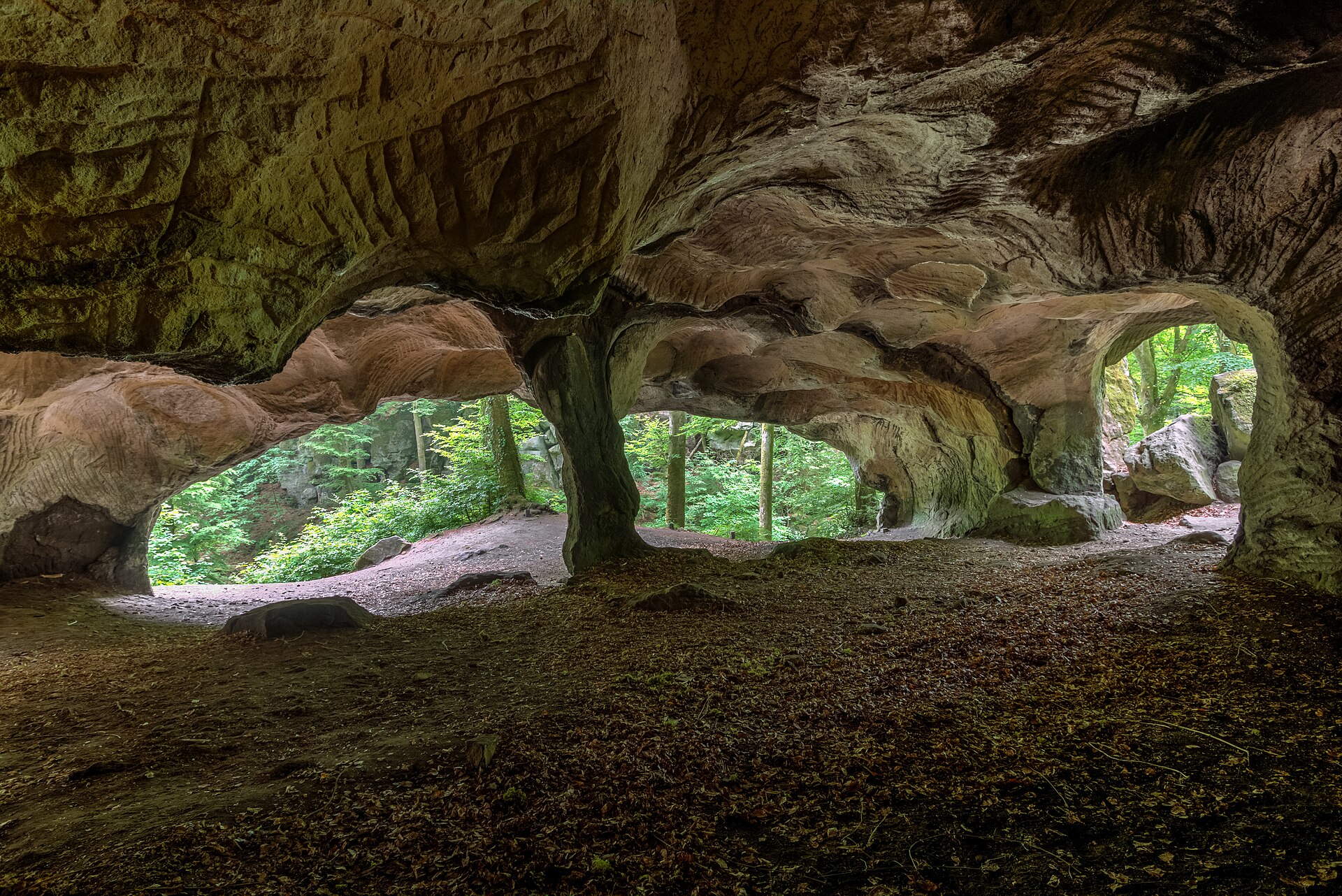 Die Felshöhle Huel Lee bei Berdorf: rötlicher Sandstein mit Abbauspuren, durch zwei Öffnungen fällt Licht vom Wald herein.