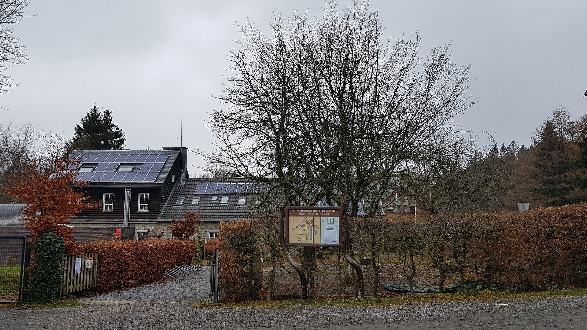 Das Naturzentrum Haus Ternell mit dunkler Holzfassade und Solardach hinter einer Hecke, davor eine Informationstafel und kahle Laubbäume.